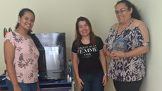 #pratodosverem: Imagem mostra coordenadora do Programa de Atenção Básica e Saúde da Família, Maria Helena Oliveira, professora Lívia Ávila e secretária de Saúde de Santos Dumont, Cláudia Paschoal. Cláudia segura um protetor facial. Atrás delas aparece a impressora 3D do Campus Santos Dumont do IF Sudeste MG, utilizada na produção. Foto: Lívia (arquivo pessoal).