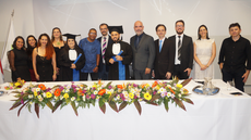 #pratodosverem: Imagem mostra formandos ao lado de componentes da mesa de honra, servidores homenageado e parte da equipe de organização da sessão solene de colação de grau. Eles estão em pé, atrás da mesa de honra da solenidade.