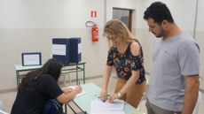 #pratodosverem: Imagem mostra eleitora sentada e registrando seu voto enquanto a bibliotecária Érica Melo e o professor Tadeu Pereira, em pé, organizam listas sobre uma mesa. No fundo, aparece uma urna de votação. A foto, de 2019, remete ao concurso para escolha do nome da Biblioteca do Campus Santos Dumont. O nome vencedor foi Acyr Loureiro Lima.