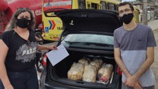 #pratodosverem: Imagem mostra representante da Sociedade São Vicente de Paulo ao lado do professor Carlos Artur Leal, do IF Sudeste MG, em entrega de cestas. Ao fundo, aparece um veículo com porta-malas aberto, que comporta algumas cestas doadas pelo projeto Educação e Cidadania. Eles estão de máscara e próximos à fachada do Campus Santos Dumont.
