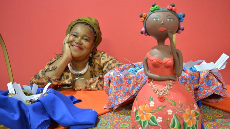 WhatsApp Image 2019-06-17 at 17.29.41.jpg À esquerda, professora Vanda Ferreira, sentada à mesa, sorri. À direita, uma boneca namoradeira está posicionada sobre a mesa.