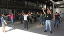 #pratodosverem: Imagem mostra professor de Educação Física Gustavo Pasqualini orientando prática de Ginástica Laboral no espaço de vivência do Bloco 1 do Campus Santos Dumont. Vários servidores fazem exercícios, com os braços projetados em diversas direções.