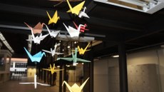 #pratodosverem: Imagem mostra móbile de tsurus feito em origami, exposto no Bloco 1 do Campus Santos Dumont. Ao fundo, aparecem o espaço de convivência, acessos a salas de aula e também o prédio da Biblioteca Acyr Loureiro Lima.