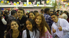 #pratodosverem: Imagem mostra estudantes do Campus Santos Dumont durante o XII Arraiá do IF, em 2022. Acima deles aparecem bandeirinhas tradicionais de festas juninas.