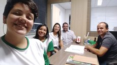 Duas alunas, uma professora e um professor e o proprietário da Empresa em reunião, sentados ao redor de uma mesa.